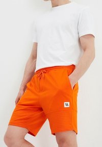 Shorts de sport orange vif avec une taille élastique et des poches latérales. Accents texturés près de l'ourlet et une petite étiquette de marque sur la jambe.