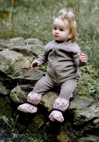 Brązowy kombinezon polarowy z przednim zamkiem błyskawicznym, ściągaczami na mankietach oraz różowymi wodoodpornymi bucikami z czarnymi paskami. Dziecko siedzi na kamieniu.