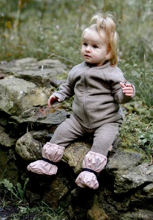 Hnědý overal z fleecu se zapínáním vpředu na zip, elastickými manžetami a růžovými voděodolnými botičkami s černými pásky. Dítě sedí na kameni.