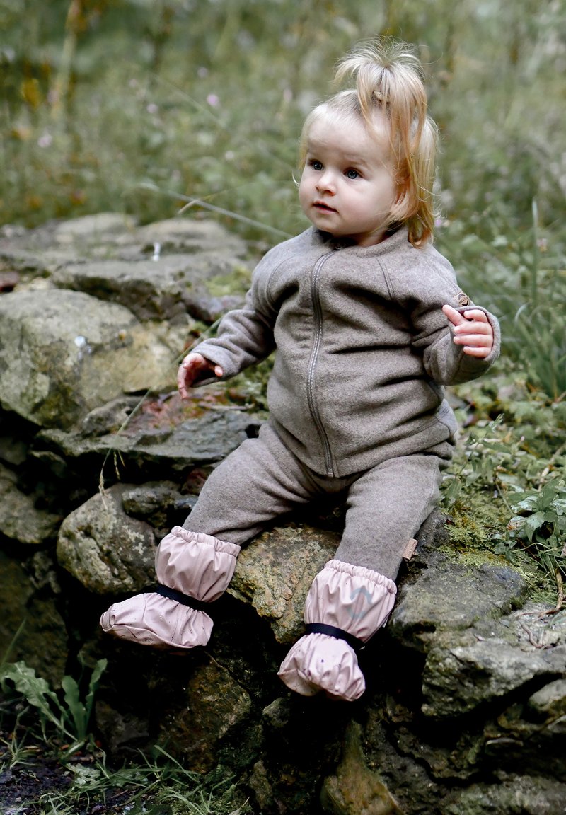 Brązowy kombinezon polarowy z przednim zamkiem błyskawicznym, ściągaczami na mankietach oraz różowymi wodoodpornymi bucikami z czarnymi paskami. Dziecko siedzi na kamieniu.