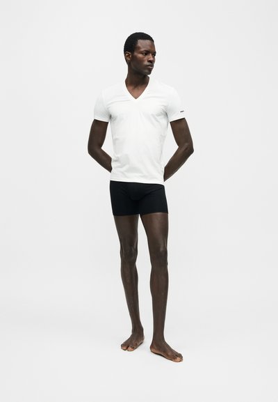 Άνδρας στέκεται ξυπόλητος, φοράει λευκό T-shirt με V-λαιμόκοψη και μαύρα boxer, κοιτώντας προς τα πλάγια σε απλό λευκό φόντο.