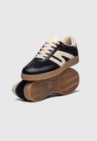 Scarpa sportiva nera e crema con tomaia in pelle e tessuto, suola in gomma testurizzata e lacci crema. Presenta il logo sul lato e accenti decorativi.