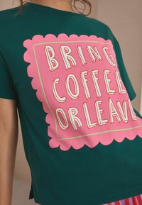 Person som bär en grön T-shirt med en stor rosa grafik som säger "TA MED KAFFE ELLER ÅK HEM."