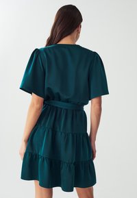 Petrolfarbenes, gestuftes Kleid aus glattem Stoff mit weitem Ausschnitt, kurzen Puffärmeln und einer taillierten Passform, die durch einen passenden Gürtel betont wird.