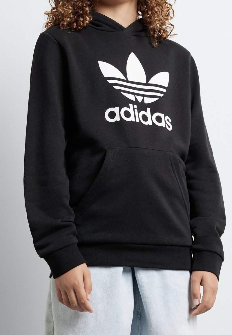 Černá mikina Adidas s velkým bílým logem na přední straně, přední kapsou a ribbenými manžetami. Materiál vypadá měkký a hladký.