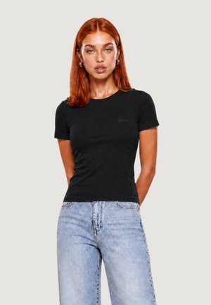Femme aux cheveux roux portant un t-shirt noir moulant et un jean bleu clair taille haute, debout devant un fond uni.
