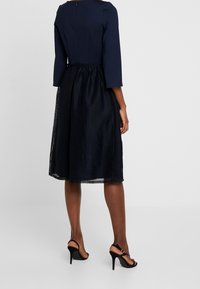Navy dress with a fitted top and a pleated mesh skirt, featuring three-quarter sleeves and a zipper closure at the back; worn with black heels.