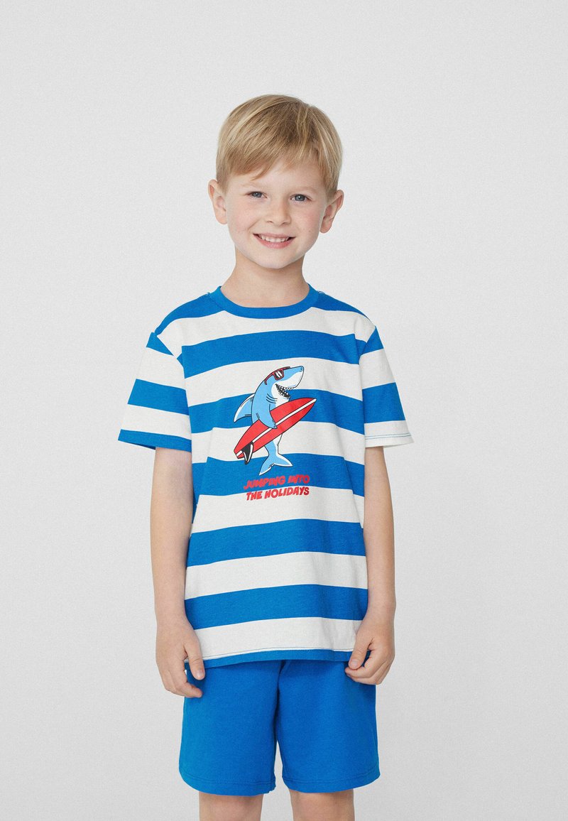 T-shirt en coton rayé bleu et blanc avec un graphique de requin et du texte rouge, associé à un short bleu uni. Texture lisse dans l'ensemble.