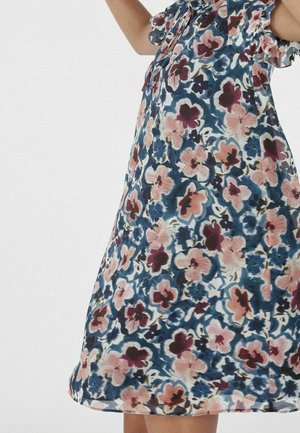 Barn iført en blå kjole med blomster i pink, lilla og hvid samt flæser på ærmerne, løfter armene mod en ensfarvet baggrund.