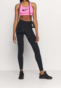 Kobieta w czarnych legginsach, różowym biustonoszu sportowym Nike, czarnych sneakersach, trzymająca czarną taśmę oporową na ramionach, na tle białej ściany.