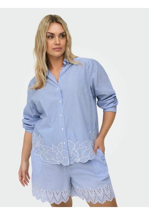Kvinde iført blå og hvidstribet skjorte med knapper med blomsterbroderi og matchende shorts med bølget blondetrim.
