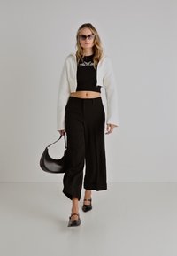 Balts saīsināts jaciņa, melna pieguļoša blūze, plata melna bikses un melnas kurpes; tur rokās maza melna soma; saulesbrilles papildina izskatu.