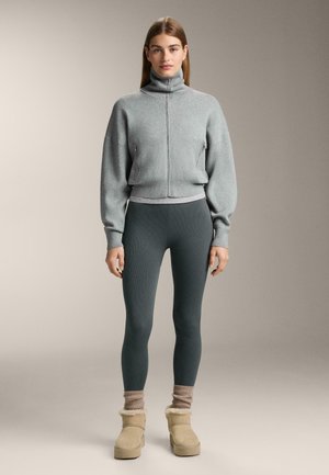 Siva zadrga majica s visokim ovratnikom, rebrastim manžetama, u kombinaciji s tamnozelenim rebrastim tajicama i bež čizmama s krznom. Prisutne su meke teksture.