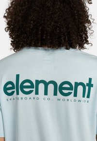 Lyseblå kortærmet t-shirt med et stort grønt "element" logo og mindre tekst, der siger "SKATEBOARD CO. WORLDWIDE" på ryggen.