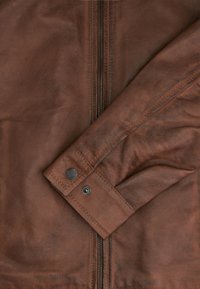 Brown leather jacket featuring a visible zipper and button detail on the cuff, with a soft, textured finish.