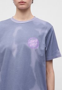 Personne portant un t-shirt lavande avec un logo circulaire « Santa Cruz » sur la poitrine gauche, debout devant un fond clair uni.