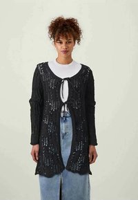 Melnas adītas kardigāna ar atvērtu priekšu, sasienamu aizdari, garām piedurknēm un mašīnas izstrādātā, mežģīņu līdzīgā rakstā. Valkāta virs baltas blūzes un gaiši zilām svārkiem.