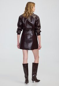 Vestido mini de cuero marrón oscuro con cintura ajustada y mangas ligeramente abullonadas. Botas de charol marrón hasta la rodilla con detalle de patrón de cocodrilo.