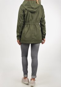 Oliivinvihreä takki, jossa on kiristysnauha vyötäröllä, huppu ja joustavat hihansuut, valmistettu sileästä kankaasta. Yhdistetty harmaisiin kapealahkeisiin farkkuihin ja vaaleisiin tennareihin.