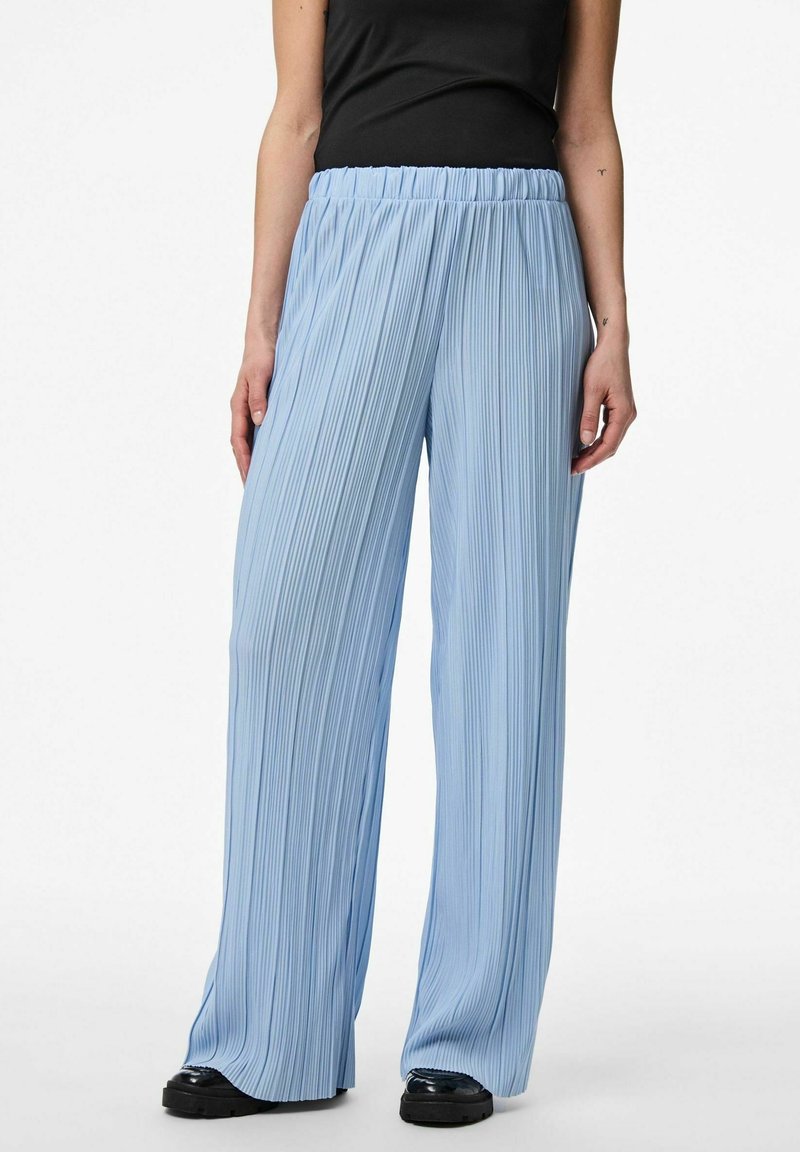Femme portant un pantalon plissé large bleu clair et des chaussures noires, avec un haut noir sans manches visible de la taille vers le bas.