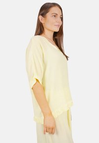 Hellgelbe Bluse mit lässigem Schnitt, kurzen Ärmeln und einem dezenten strukturierten Muster. Verfügt über einen V-Ausschnitt und einen rüschierten Saum.