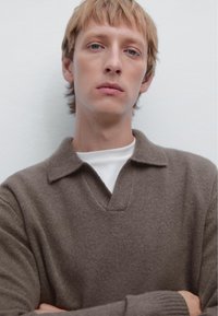 Brown, v-neck sweater with a textured knit, featuring a collar and layered over a white shirt. Model has short hair and is standing against a light wall.