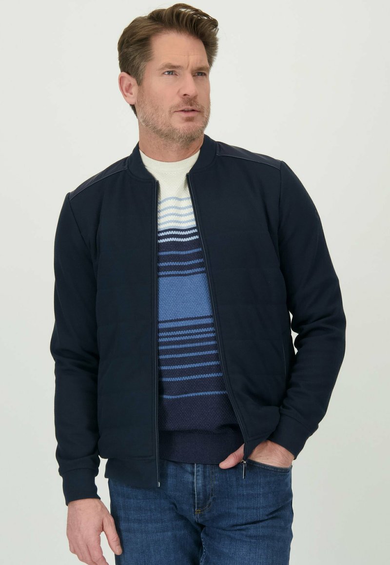 Marineblauwe zip-up jack met een geribbelde kraag en schouderpatches; gelaagd over een gestreepte trui in blauw en wit; gecombineerd met blauwe jeans.