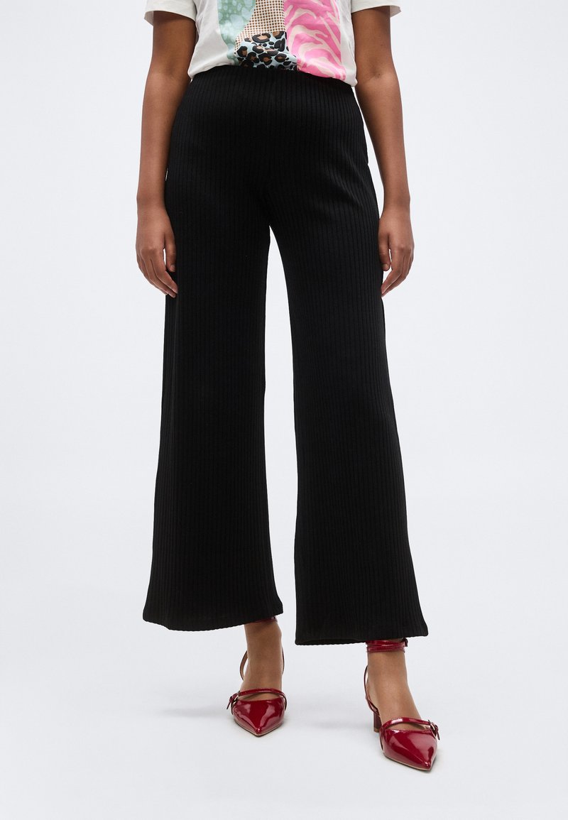 Άτομο που φοράει μαύρο ριμπ παντελόνι με φαρδιά μπατζάκια και κόκκινα ψηλοτάκουνα παπούτσια με μυτερή μύτη, στέκεται μπροστά σε ένα απλό ανοιχτό φόντο.