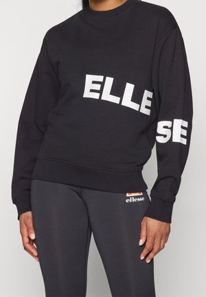 Osoba nosząca czarną bluzę i spodnie z białym napisem logo "ellesse" na piersi, rękawie i górnej części uda.