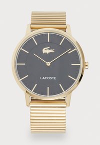 Apvalus auksinės spalvos laikrodis su tekstūruotu auksiniu metaliniu apyrankiu, juodu ciferblatu, auksinėmis detalėmis ir Lacoste logotipu, esančiu virš prekės pavadinimo.