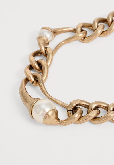 Bracelet à chaîne en or épaisse avec deux grandes perles blanches serties dans des maillons irréguliers de forme organique sur un fond clair uni.