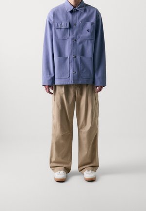 Personne portant une veste bleue boutonnée avec quatre poches avant, un pantalon beige à jambes larges et des baskets blanches, debout devant un fond uni.
