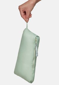 Main tenant une pochette zippée vert clair par sa boucle en tissu contre un fond blanc.