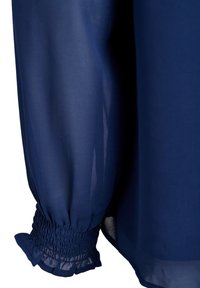 Blouse en voile bleu marine avec des poignets froncés, une silhouette fluide et de subtils détails à volants aux bords des manches. Texture lisse.