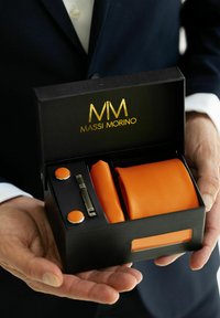 Corbata naranja, gemelos naranjas y un sujetacorbata plateado en una caja negra con logotipo dorado. La caja es sostenida en una mano, mostrando claramente su contenido.