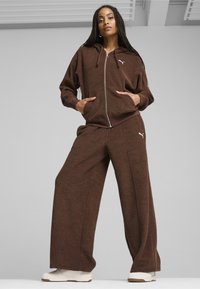 Hnedá mikina s kapucňou na zips s jemnou textúrou a klokankovými vreckami, doplnená zodpovedajúcimi širokými nohavicami. Obe obsahujú nenápadný detail loga.