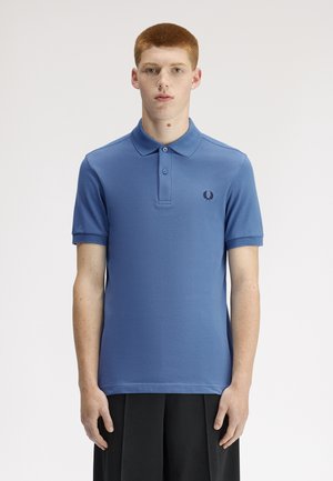 Ung mand med kort rødt hår iført en blå polo med en lille mørk logo, stående foran en ensfarvet lys baggrund.