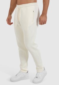 Cremfarbene Jogginghosen aus weichem Stoff, ausgestattet mit einem elastischen Bund mit Kordelzug, seitlichen Taschen und subtiler Markenkennzeichnung auf der Vorderseite.