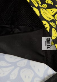 Černý lem s hladkým povrchem, barevný vzor žlutých usmívajících se tváří na vnější tkanině a štítek označený "SMILEY ORIGINALS."