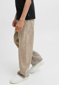 Bež traperice sproščene kroja z zbledelim videzom, vidnimi šivi in stranskimi žepi, v kombinaciji z belimi tenisicami.