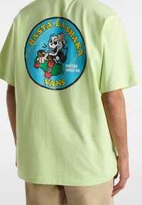 Camiseta de algodón verde claro con un gráfico circular de un esqueleto en una patineta, con los textos "HASTA LA SHAKA" y "VANS" en colores vivos.