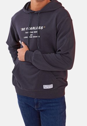 Hombre con sudadera con capucha gris oscuro con bolsillo delantero y texto "BE FEARLESS TAKE THE RISK OR LOSE THE CHANCE", combinado con jeans azules.