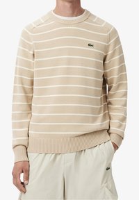 Beige svetr s bílými vodorovnými pruhy, žebrovaný límec a manžety, vyrobený z měkkého materiálu. Na hrudi je zelené logo Lacoste.