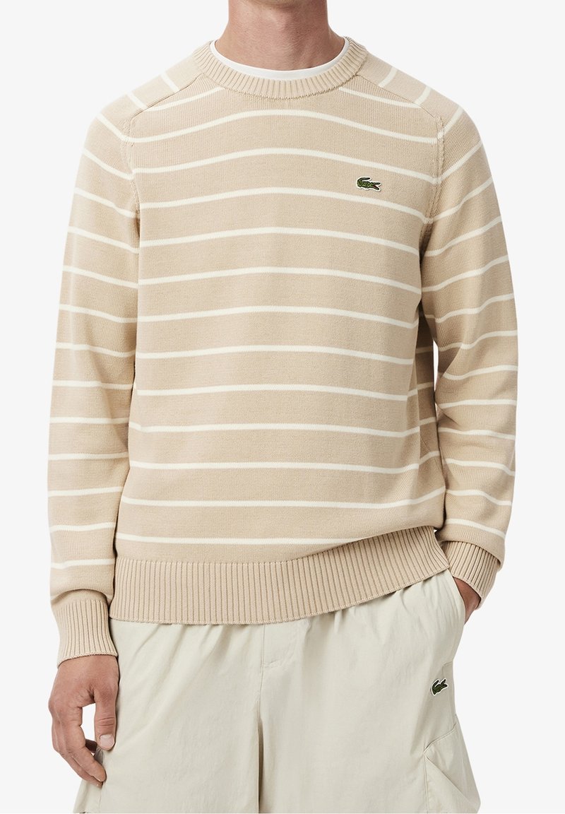 Beige svetr s bílými vodorovnými pruhy, žebrovaný límec a manžety, vyrobený z měkkého materiálu. Na hrudi je zelené logo Lacoste.