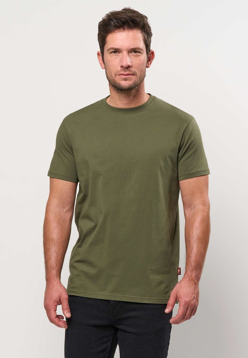Homme aux cheveux courts et bruns portant un t-shirt à manches courtes vert olive et un pantalon noir, debout devant un fond clair uni.