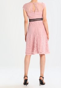 Rosa spetsklänning med korta ärmar, en urklippt rygg och en svart spetsdetalj i midjan. Mjuk textur och knä-lång nederdel. Bärs med svarta klacksandaler.