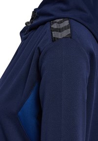 Donkerblauwe, gestructureerde jas mouw met een blauwe diamantvormige patch en een zwart-wit patroon stofdetail vlakbij de schouder.