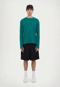 Hosszú ujjú türkizkék ing varrott díszítésekkel, fekete cargo rövidnadrággal, oldalzsebekkel, és fehér sportcipővel párosítva. Fekete sapka a fejen.