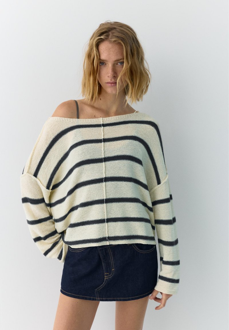 Jeune femme aux cheveux blonds mi-longs porte un pull crème à épaules dénudées avec des rayures noires et une mini-jupe en denim foncé.