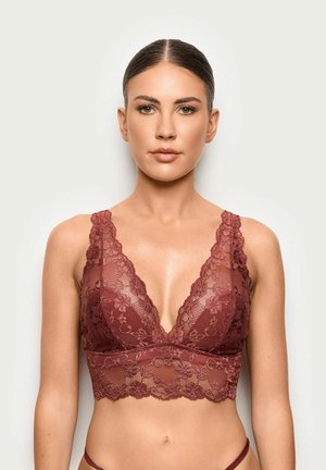 Frau mit zurückgebundenem braunem Haar trägt ein tiefrotes Spitzen-Bralette und steht vor einem schlichten hellen Hintergrund, nach vorne blickend.